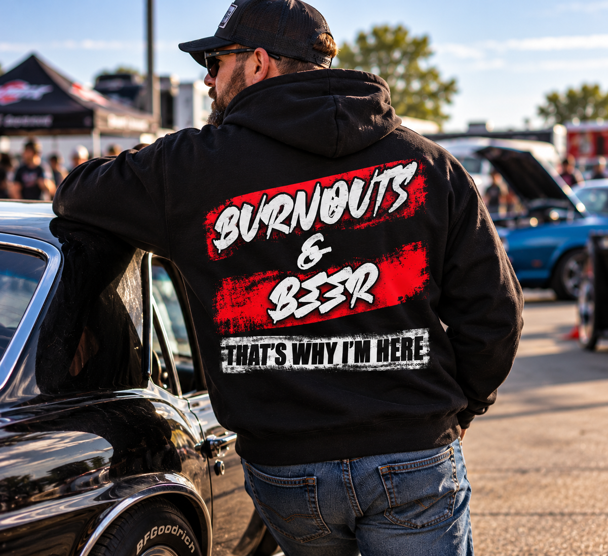 Man wearing black Burnouts & Beer racing hoodie leaning against a classic muscle car at an automotive event
