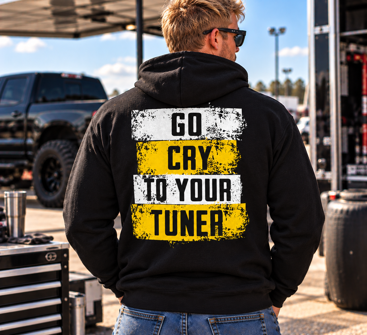 Blonde man wearing a black Car Chix Go Cry to Your Tuner hoodie featuring distressed white and yellow racing graphics on the back at a motorsports event.