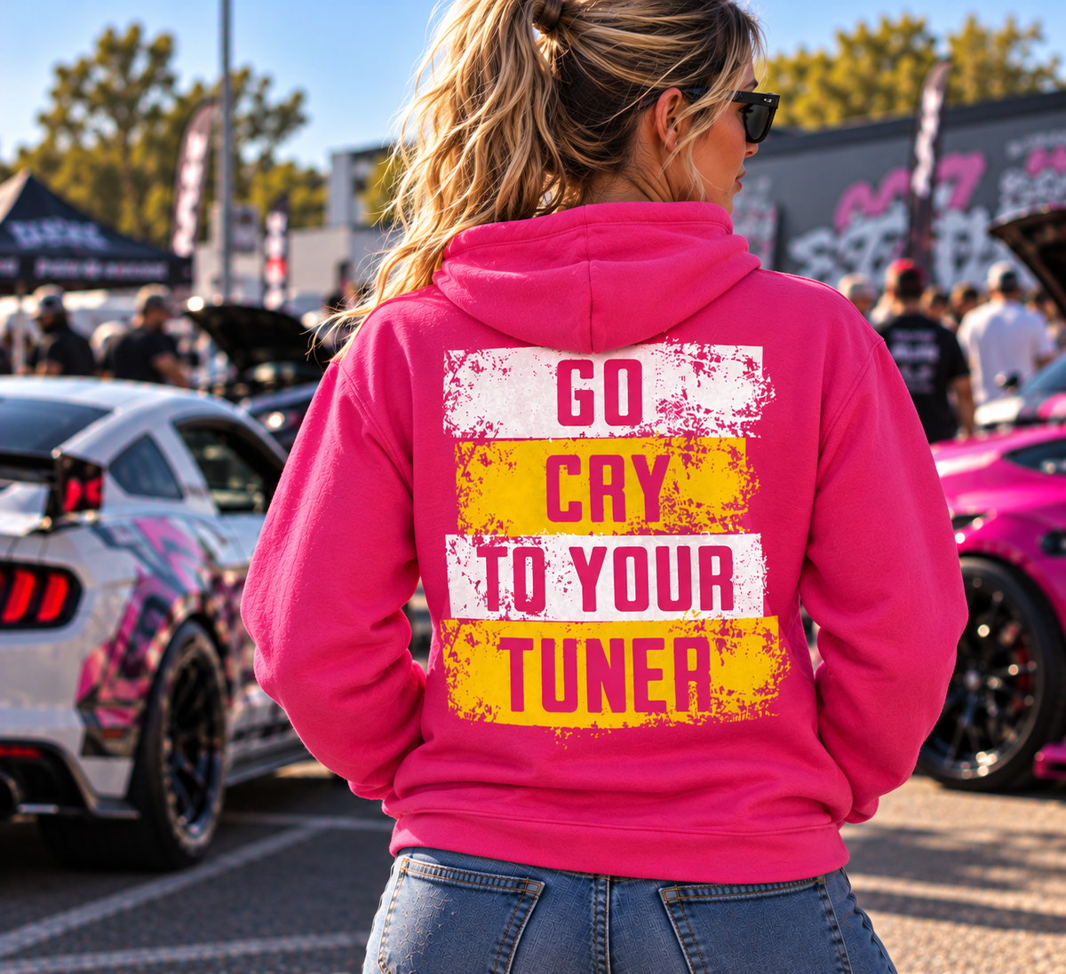 Woman wearing a dark pink Car Chix Go Cry to Your Tuner hoodie with bold white and yellow distressed graphics and pink knockout lettering at an outdoor car show.