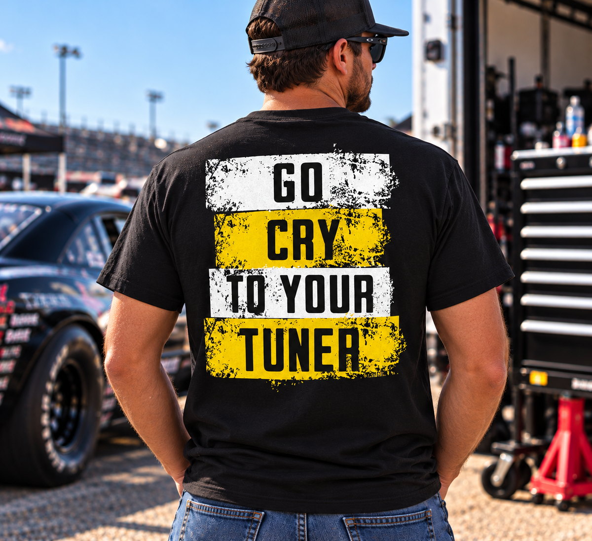 Man wearing a black Car Chix Go Cry to Your Tuner T-shirt featuring distressed white and yellow racing graphics on the back at a motorsports event.