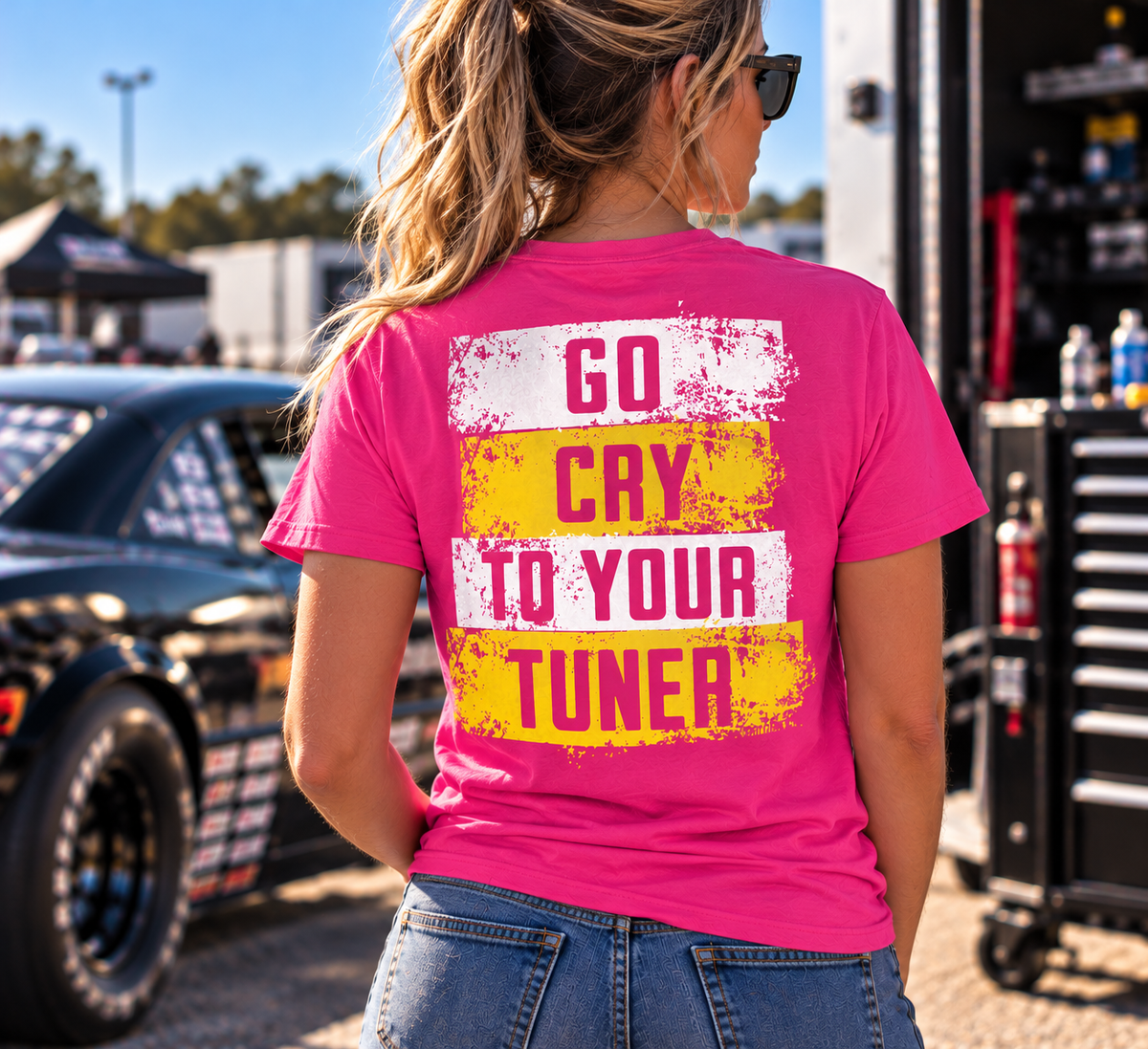 Woman wearing a dark pink Car Chix Go Cry to Your Tuner T-shirt featuring a distressed white and yellow racing design on the back at a drag strip.