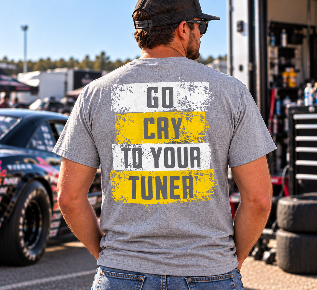 Man modeling a sport grey Car Chix Go Cry to Your Tuner T-shirt with white and yellow distressed back graphic in a motorsports pit area.