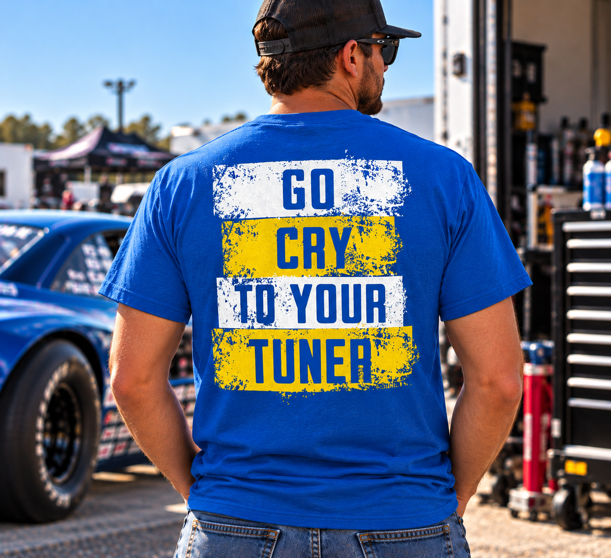 Man wearing a royal blue Go Cry to Your Tuner T-shirt by Car Chix with bold white and yellow distressed lettering at a drag racing event.