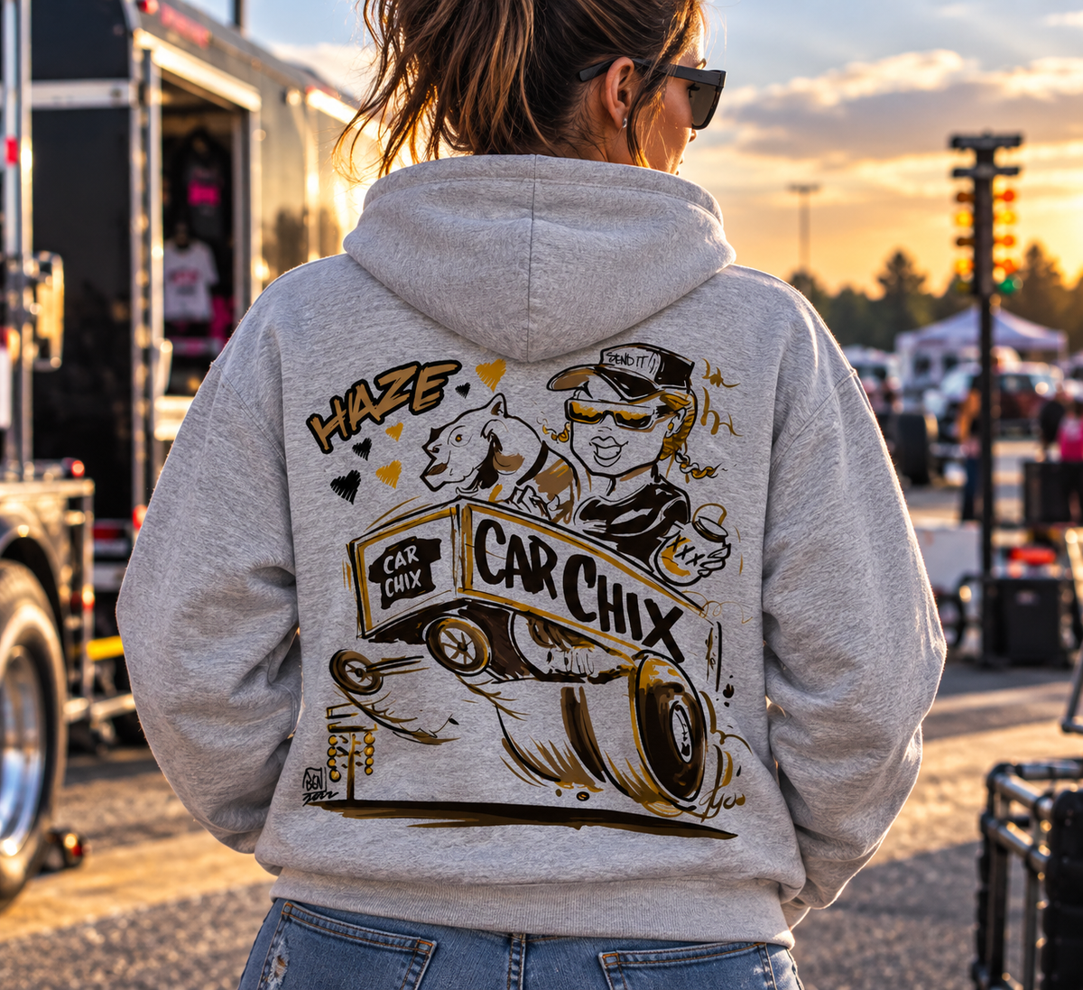 Woman wearing grey Car Chix Haze Memorial Hoodie featuring original Haze and Car Chix drag racing artwork on the back at a racetrack