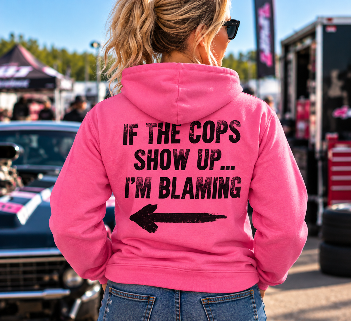 Woman wearing pink Car Chix If The Cops Show Up I'm Blaming hoodie with black distressed lettering and left-pointing arrow on the back at a racing event.