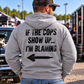 Man wearing grey Car Chix If The Cops Show Up I'm Blaming hoodie featuring black distressed lettering and a left-pointing arrow on the back at a motorsports event.