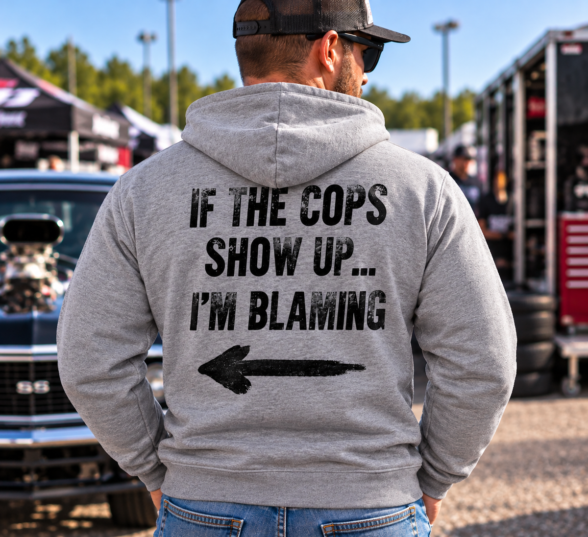 Man wearing grey Car Chix If The Cops Show Up I'm Blaming hoodie featuring black distressed lettering and a left-pointing arrow on the back at a motorsports event.