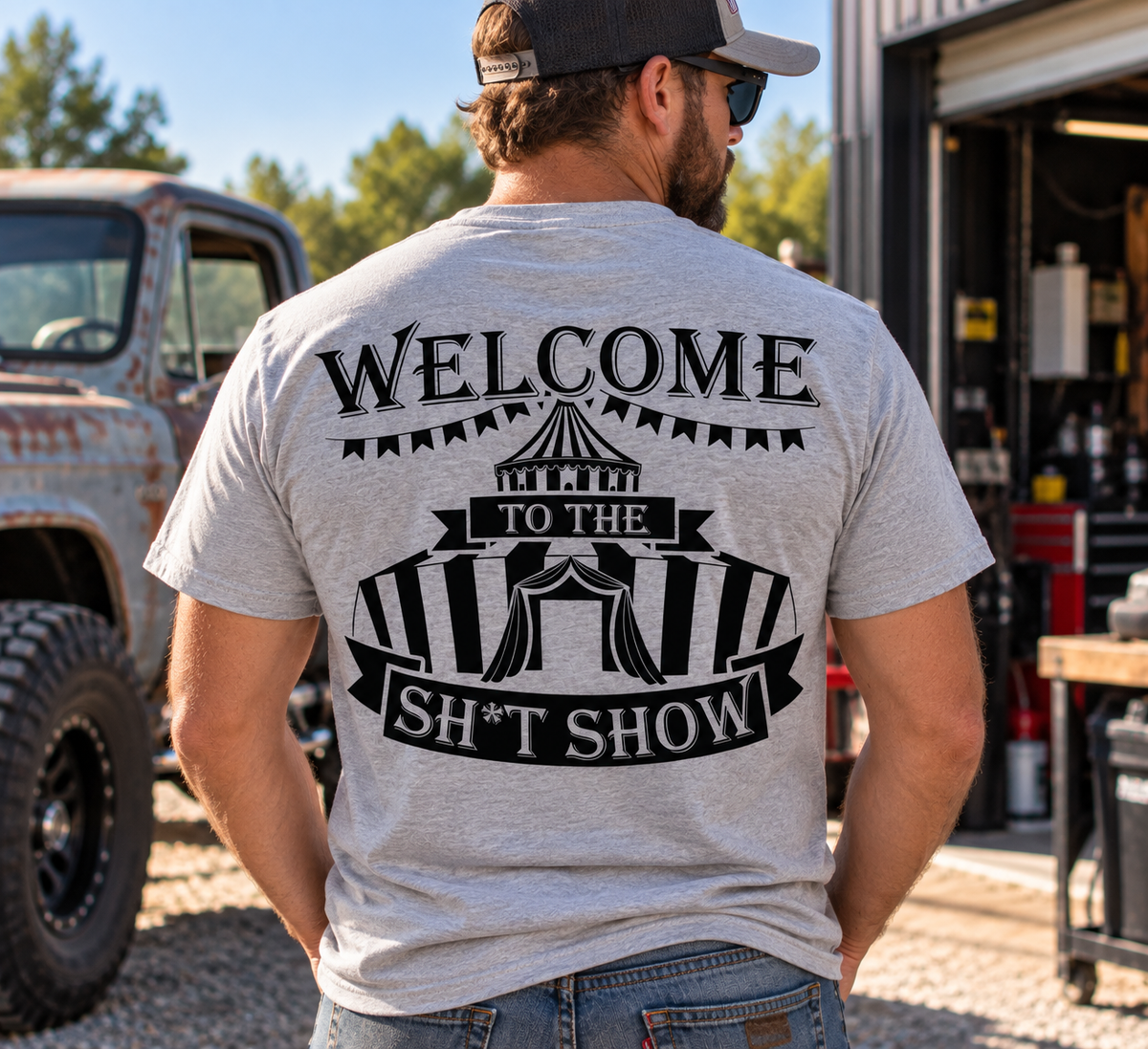 Man wearing grey Car Chix Welcome to the Sh*tshow T-shirt featuring black circus tent artwork on the back outside an automotive garage