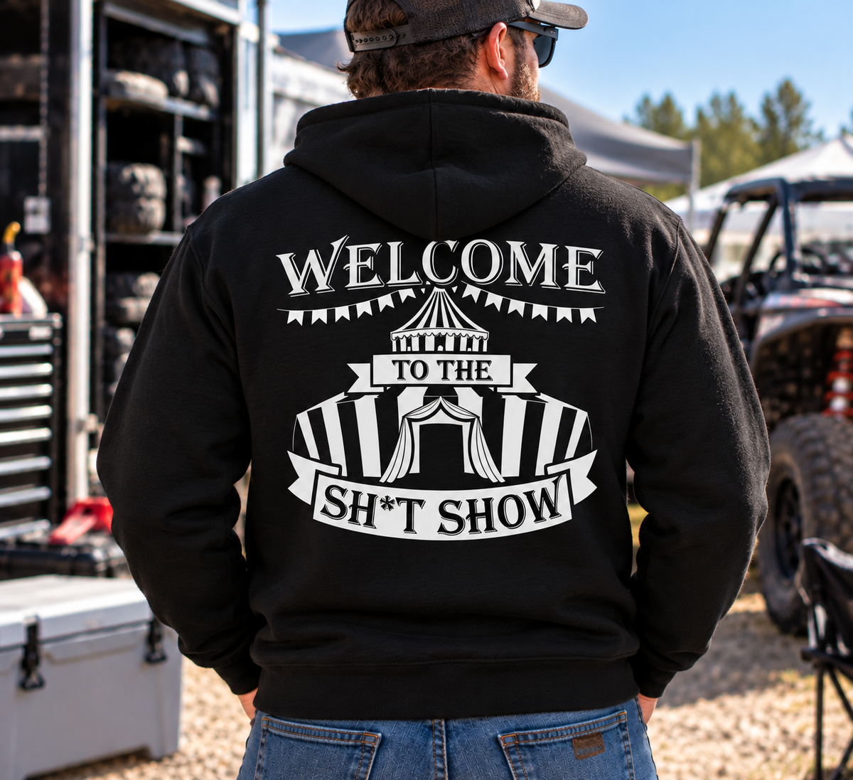 Man wearing black Car Chix Welcome to the Sh*tshow hoodie with large white circus graphic on the back at a motorsports event.