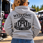 Woman wearing grey Car Chix Welcome to the Sh*tshow hoodie with large black circus-inspired back graphic at an off-road motorsports event.