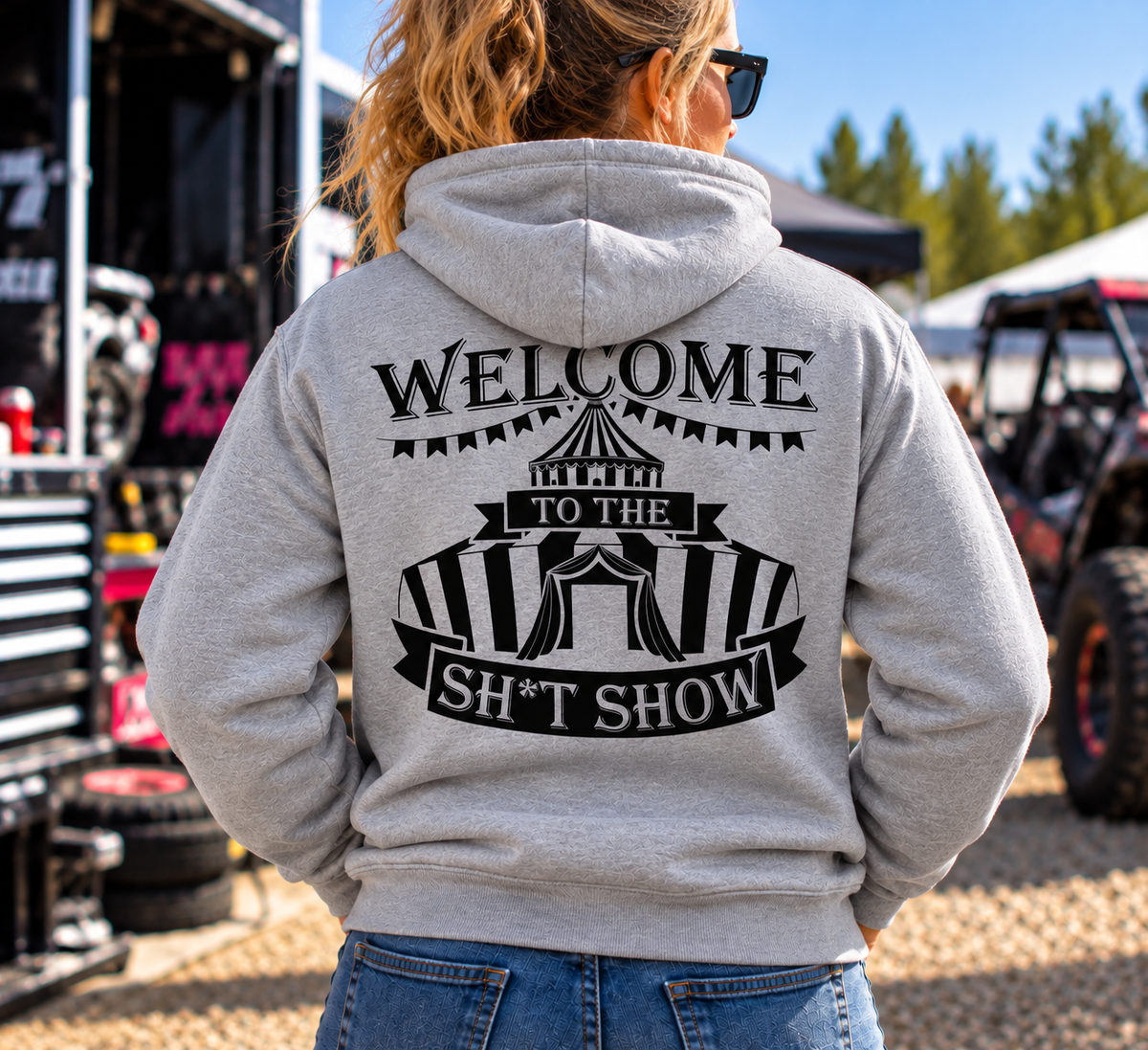 Woman wearing grey Car Chix Welcome to the Sh*tshow hoodie with large black circus-inspired back graphic at an off-road motorsports event.
