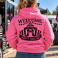 Woman wearing pink Car Chix Welcome to the Sh*tshow hoodie featuring black carnival tent artwork on the back at a racing event.