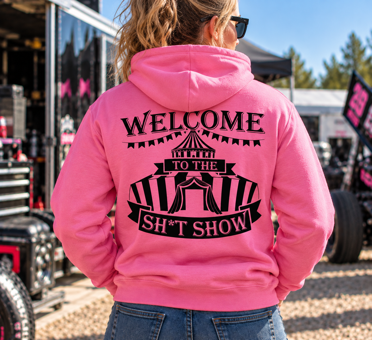 Woman wearing pink Car Chix Welcome to the Sh*tshow hoodie featuring black carnival tent artwork on the back at a racing event.