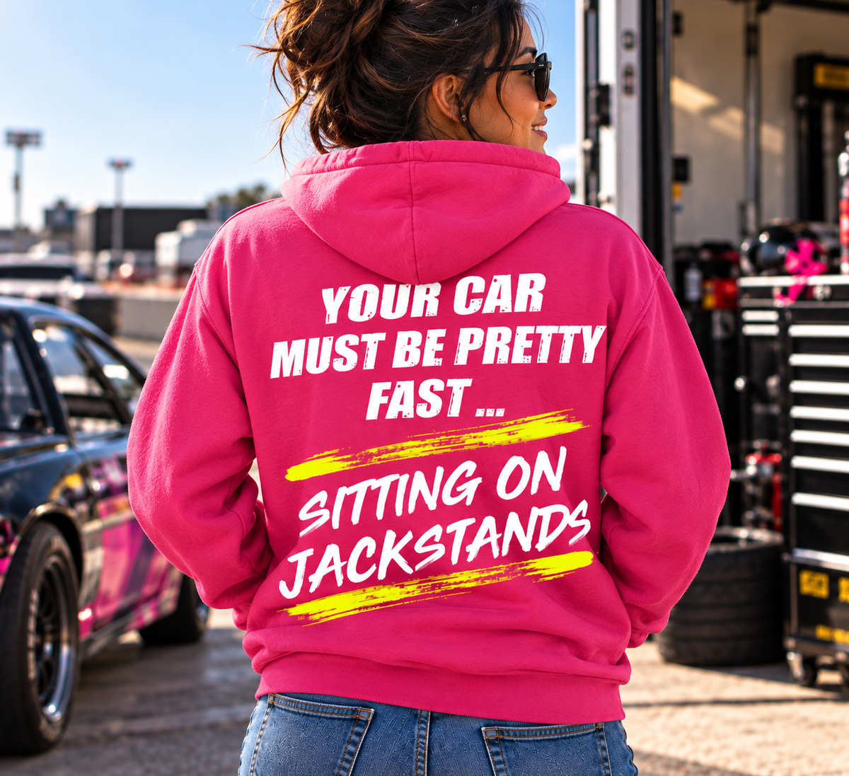 Woman wearing a dark pink Car Chix Your Car Must Be Pretty Fast Sitting on Jackstands hoodie with bold white lettering and neon yellow racing-inspired accents in a motorsports pit area.