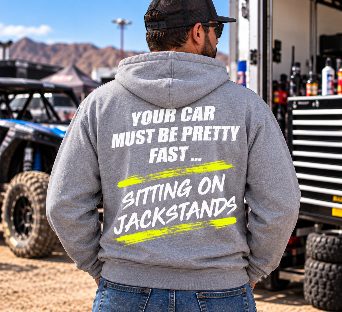 Man wearing a sport grey Car Chix Your Car Must Be Pretty Fast Sitting on Jackstands hoodie featuring white distressed text and neon yellow brushstroke graphics at an off-road motorsports event.
