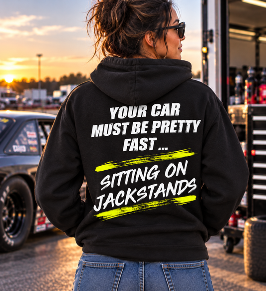 Woman wearing a black Car Chix Your Car Must Be Pretty Fast Sitting on Jackstands hoodie with distressed white lettering and neon yellow accents at a drag racing track.