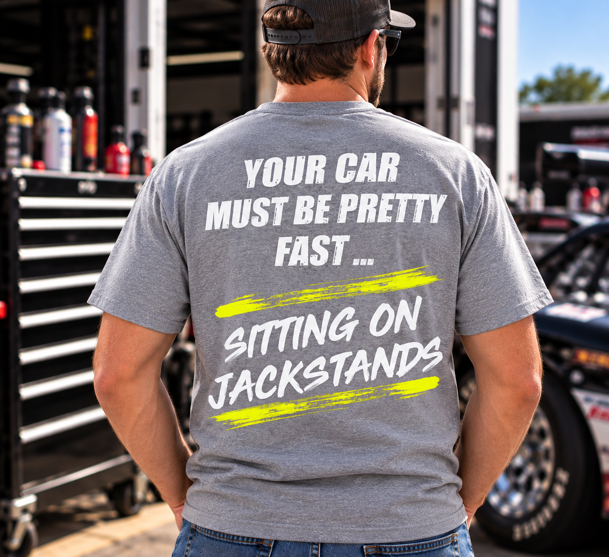 Man wearing a sport grey Car Chix Your Car Must Be Pretty Fast Sitting on Jackstands T-shirt featuring white lettering and neon yellow brushstroke graphics at a motorsports garage.