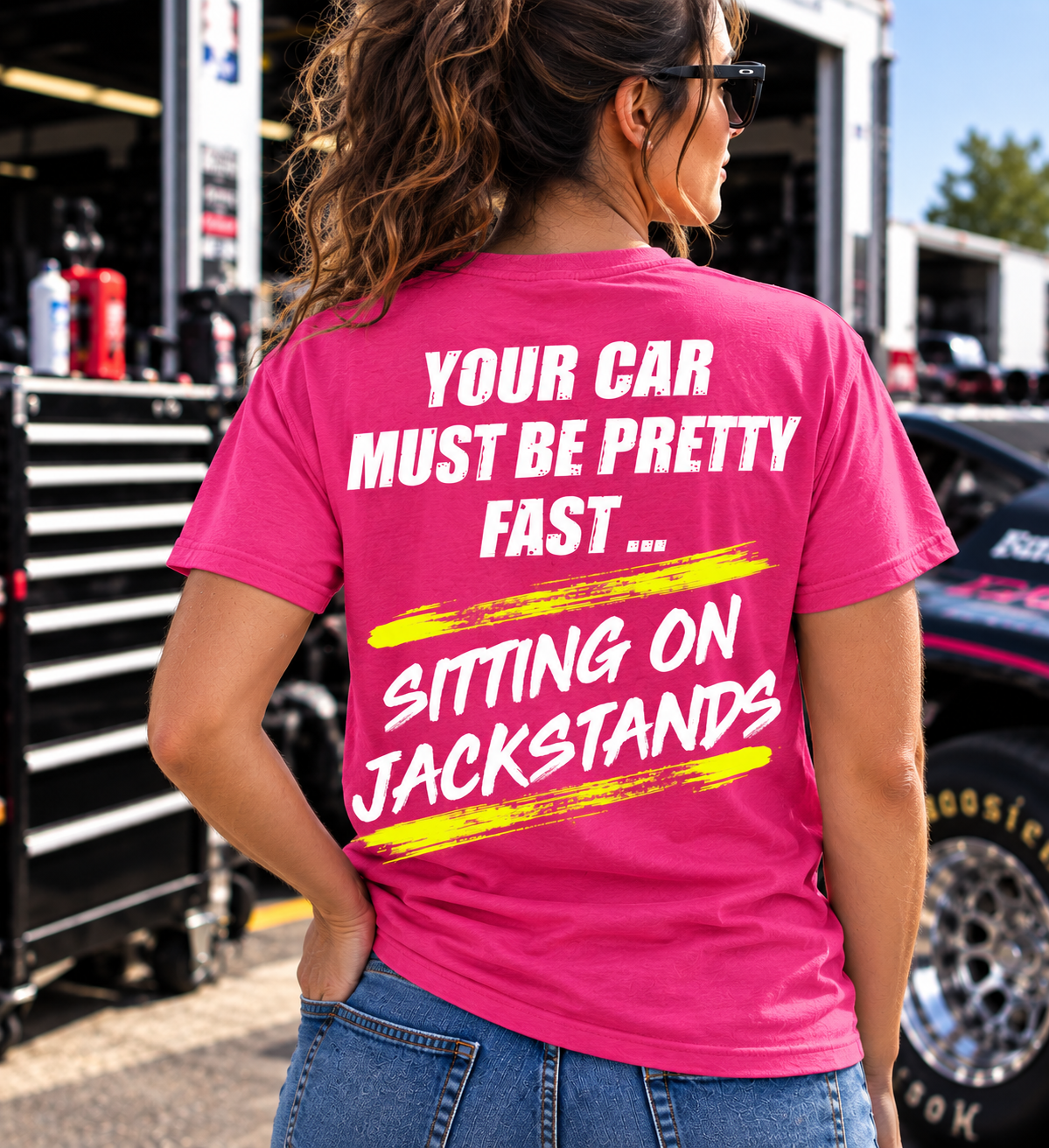 Woman wearing a dark pink Car Chix Your Car Must Be Pretty Fast Sitting on Jackstands T-shirt with white lettering and neon yellow brushstroke accents at a racetrack.