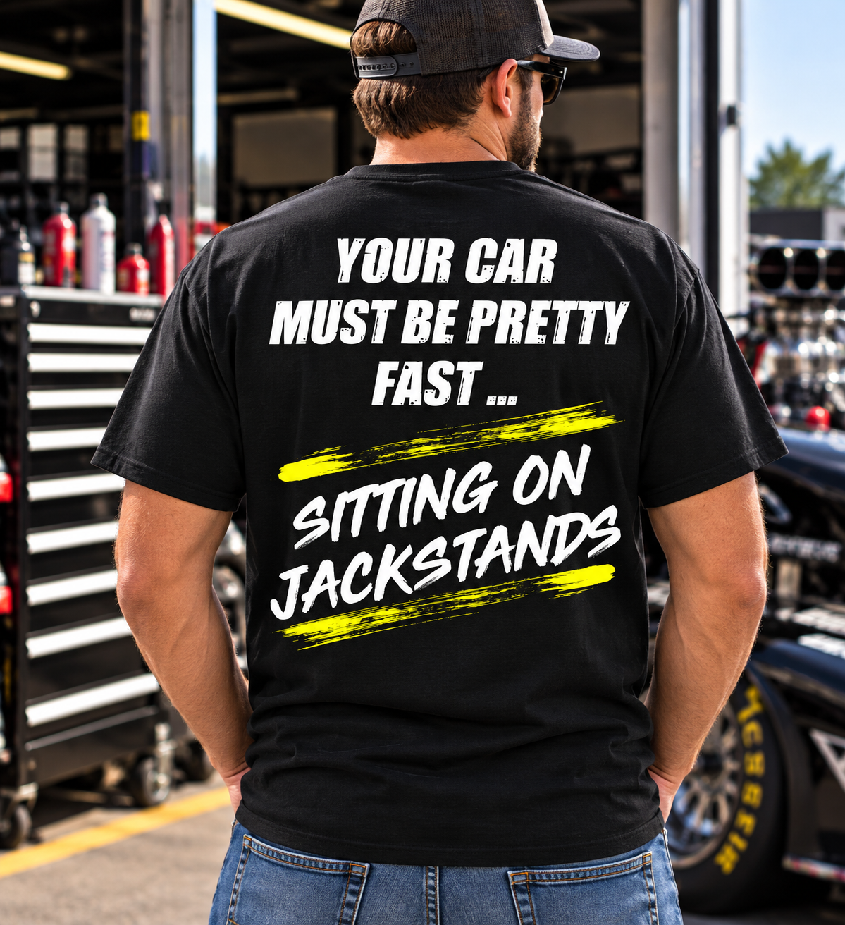 Man wearing a black Car Chix Your Car Must Be Pretty Fast Sitting on Jackstands T-shirt with white distressed lettering and neon yellow accents in a racing garage.