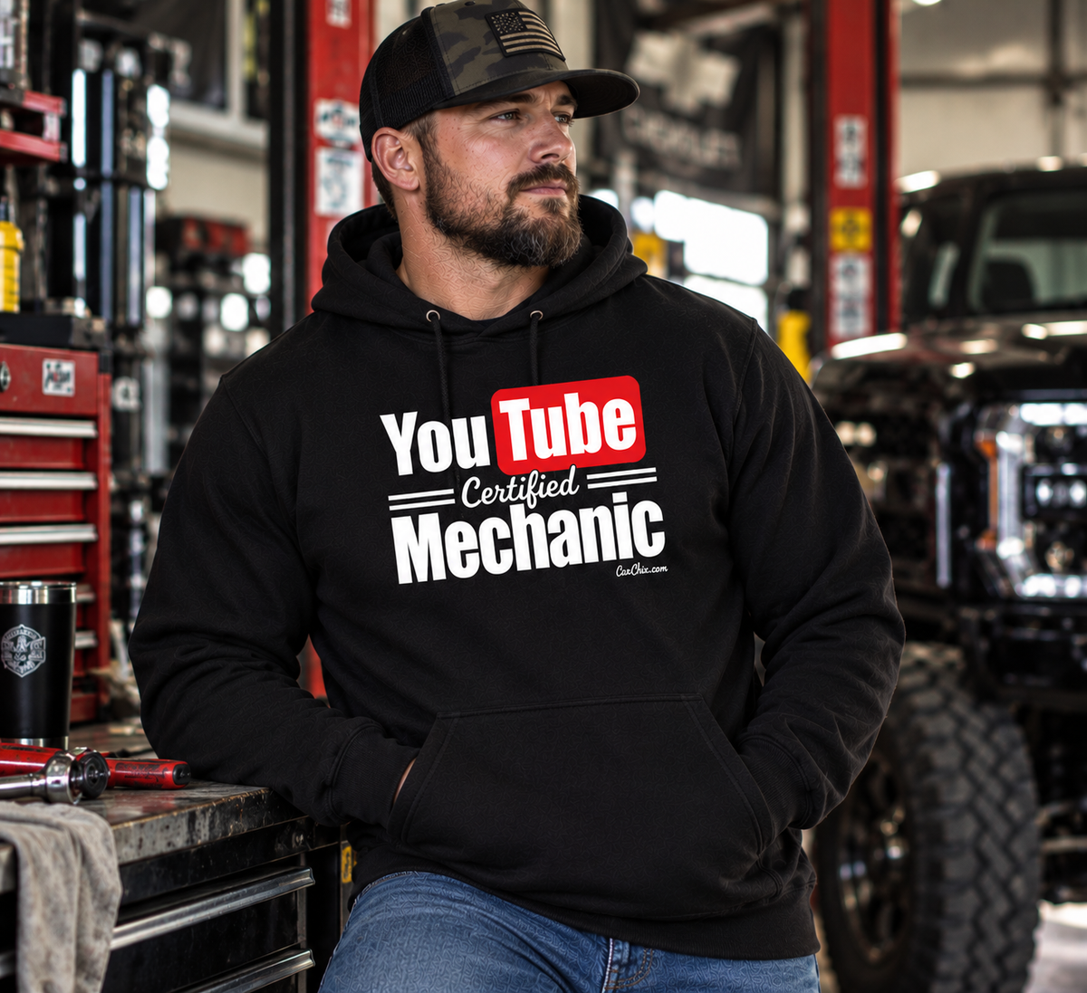 Man wearing black YouTube Certified Mechanic hoodie in an automotive garage with lifted truck and tools by Car Chix