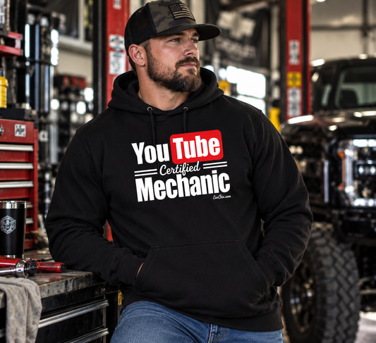 Man wearing black YouTube Certified Mechanic hoodie in an automotive garage with lifted truck and tools by Car Chix