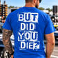 A man viewed from behind wearing a bright blue t-shirt with a distressed white graphic text that reads 'BUT DID YOU DIE?'. The background shows a blurred outdoor street scene with cars and traffic lights.