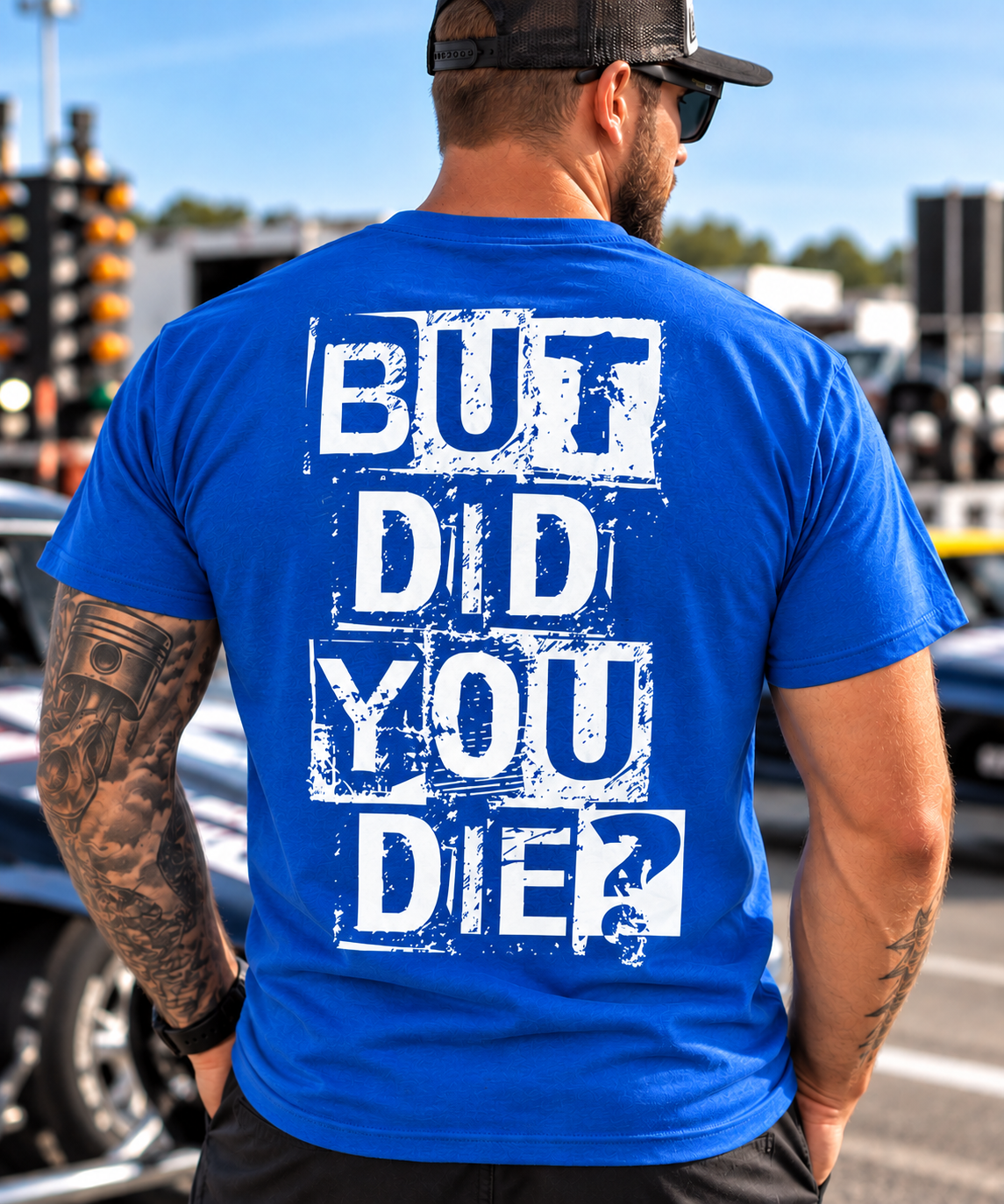 A man viewed from behind wearing a bright blue t-shirt with a distressed white graphic text that reads 'BUT DID YOU DIE?'. The background shows a blurred outdoor street scene with cars and traffic lights.