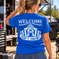 A woman wearing a bright blue t-shirt with a white graphic print on the back that reads 'WELCOME TO THE SH*T SHOW' under a circus tent design, photographed outdoors at an event.
