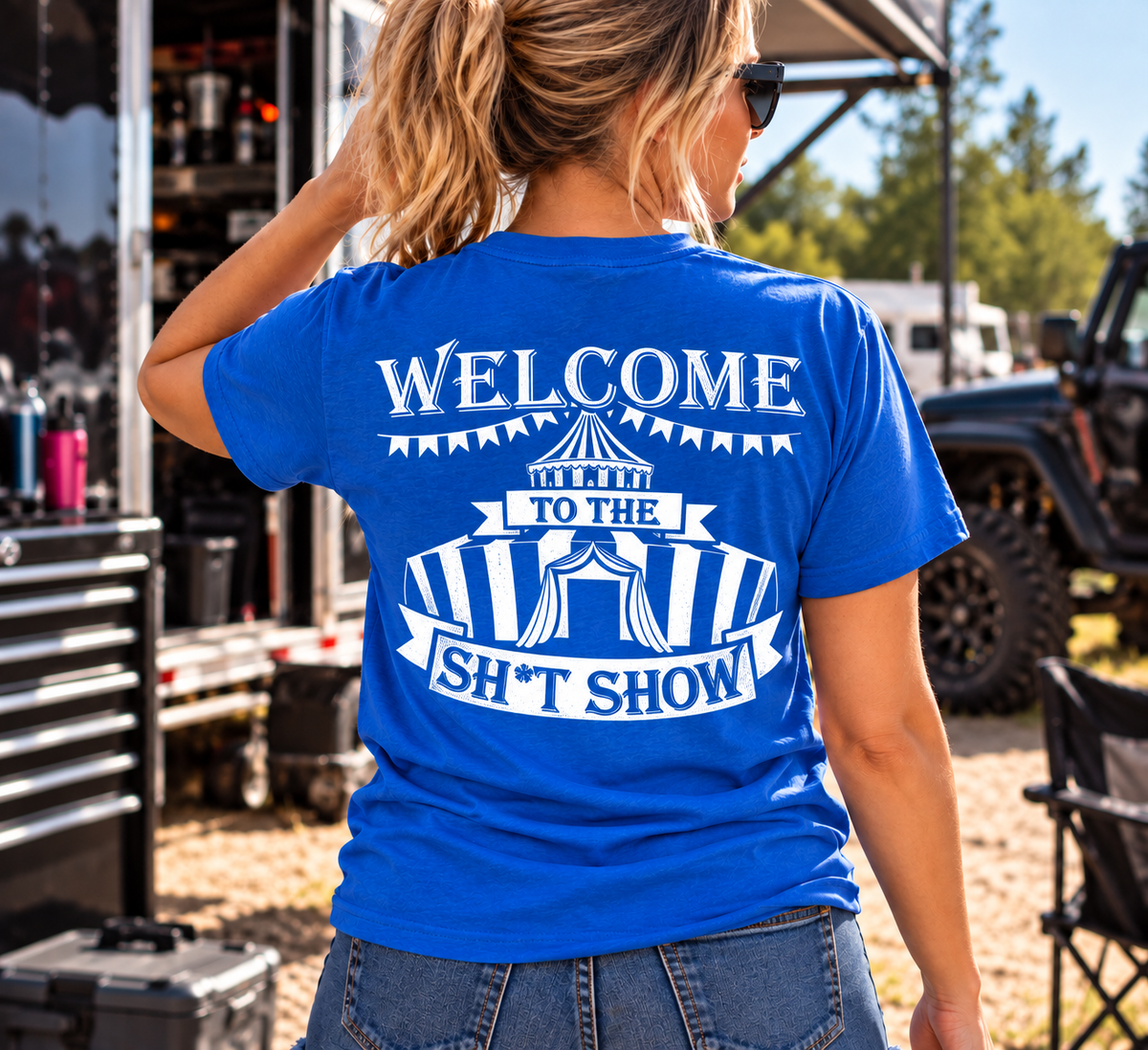 A woman wearing a bright blue t-shirt with a white graphic print on the back that reads 'WELCOME TO THE SH*T SHOW' under a circus tent design, photographed outdoors at an event.
