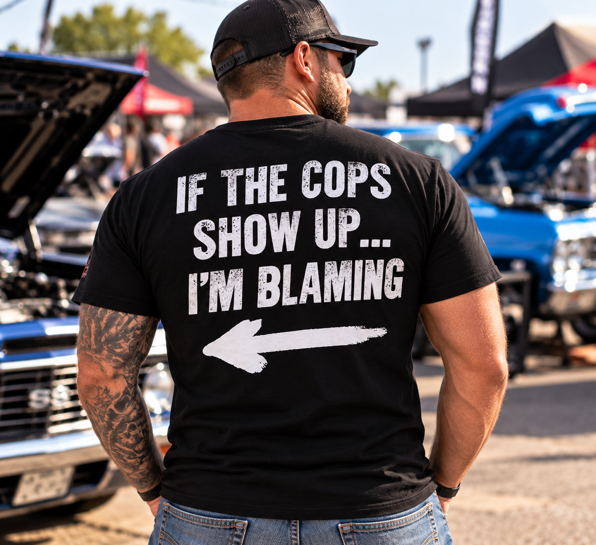 Man wearing black Car Chix If The Cops Show Up I'm Blaming T-shirt with white lettering and pointing arrow on the back at a car show.