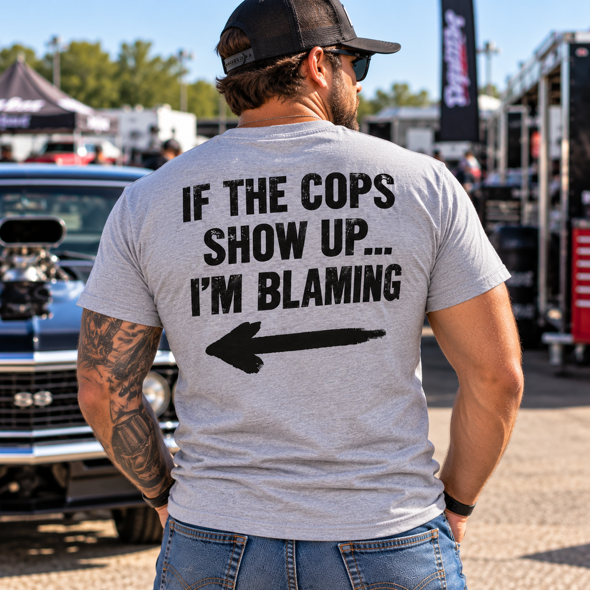 Man wearing grey Car Chix If The Cops Show Up I'm Blaming T-shirt featuring black lettering and a large pointing arrow on the back at an automotive event.
