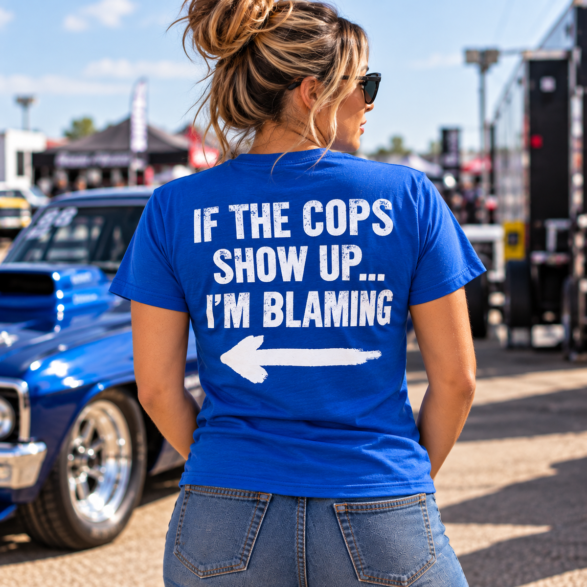 Woman wearing blue Car Chix If The Cops Show Up I'm Blaming T-shirt with white distressed lettering and pointing arrow on the back at a racing event.