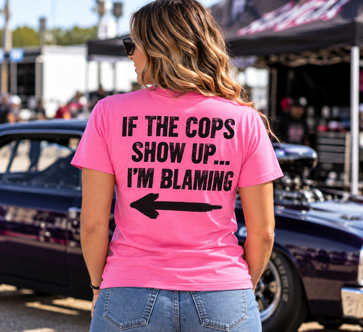 Woman wearing pink Car Chix If The Cops Show Up I'm Blaming T-shirt with distressed black lettering and left-pointing arrow on the back at a motorsports event.
