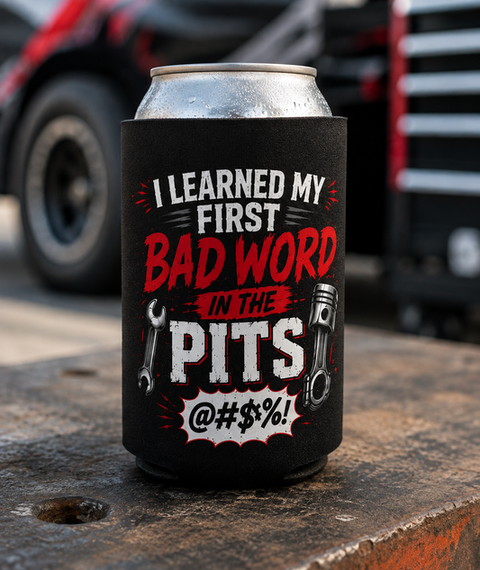 A black can cooler sleeve on a silver aluminum can, featuring the text 'I LEARNED MY FIRST BAD WORD IN THE PITS @#$$%!' with wrench and piston graphics, set on a rusty metal surface with a blurred truck in the background.