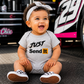 A baby wearing a light gray short-sleeve bodysuit with the text 'JUST Send It' in black and yellow, sitting on asphalt in front of a race car and tool box. The baby is also wearing a white bow headband and checkered shoes.
