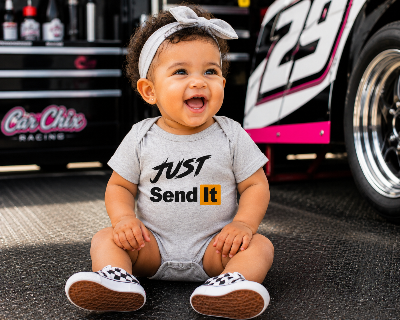 A baby wearing a light gray short-sleeve bodysuit with the text 'JUST Send It' in black and yellow, sitting on asphalt in front of a race car and tool box. The baby is also wearing a white bow headband and checkered shoes.