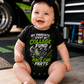 Baby wearing College Fund Race Car Parts infant racing bodysuit in a motorsports garage