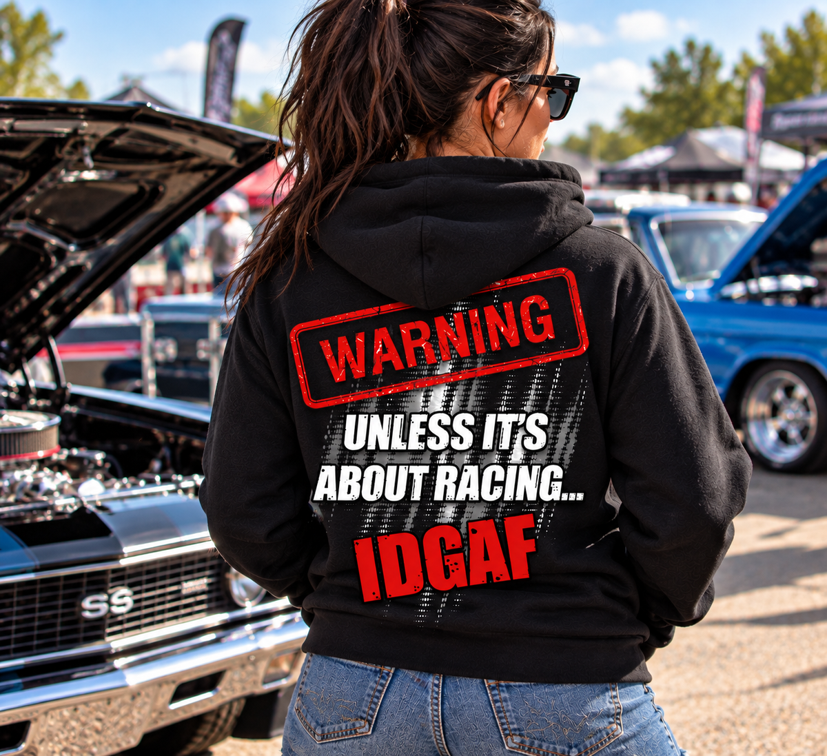 Woman wearing black Car Chix Unless It's About Racing IDGAF hoodie with bold red and white back graphic at a classic car show featuring muscle cars.