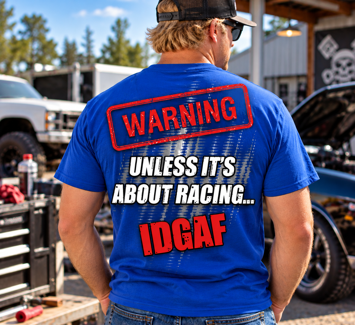 Blonde man wearing blue Car Chix Unless It's About Racing IDGAF T-shirt with bold red and white back graphic at an automotive garage.