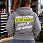 Woman wearing a sport grey Car Chix What Happens in the Garage Stays in the Garage hoodie with white and neon yellow back print, standing beside a race car in the pits.