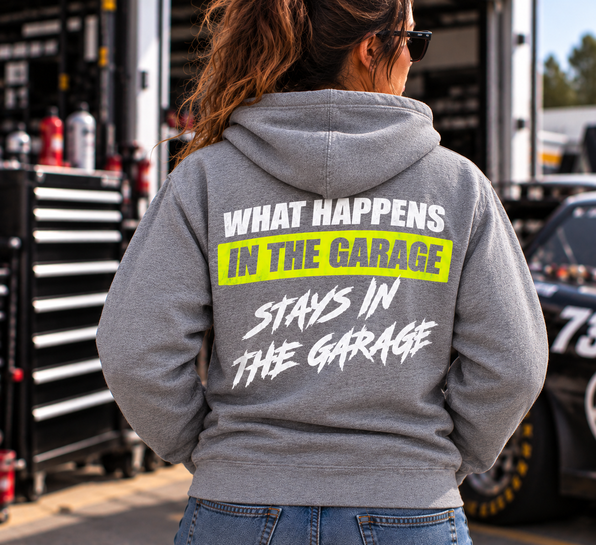 Woman wearing a sport grey Car Chix What Happens in the Garage Stays in the Garage hoodie with white and neon yellow back print, standing beside a race car in the pits.
