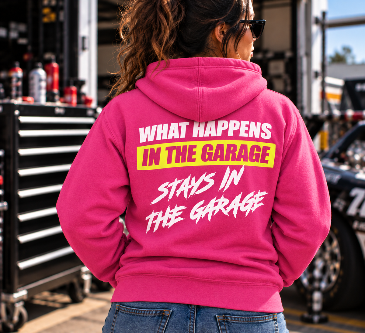 Woman wearing a dark pink Car Chix What Happens in the Garage Stays in the Garage hoodie featuring white brushstroke lettering and a neon yellow back graphic at an automotive racing garage.