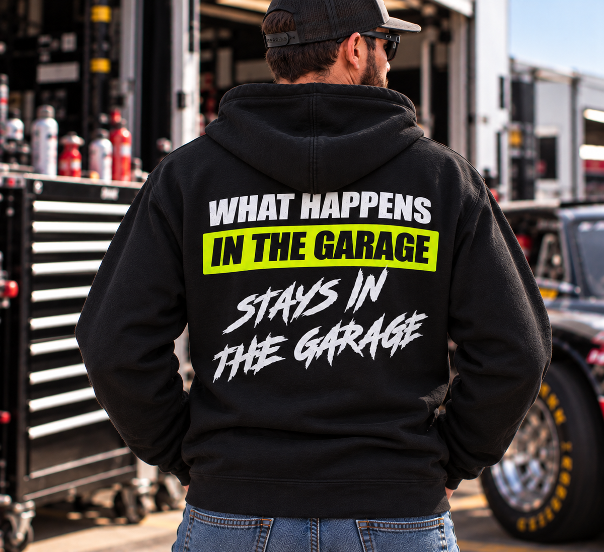 Man wearing a black Car Chix What Happens in the Garage Stays in the Garage hoodie with white lettering and neon yellow back graphic, standing in a motorsports garage.