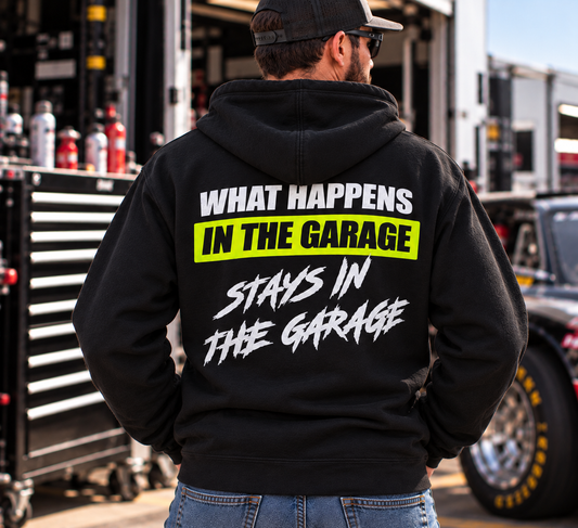 Man wearing a black Car Chix What Happens in the Garage Stays in the Garage hoodie with white lettering and neon yellow back graphic, standing in a motorsports garage.