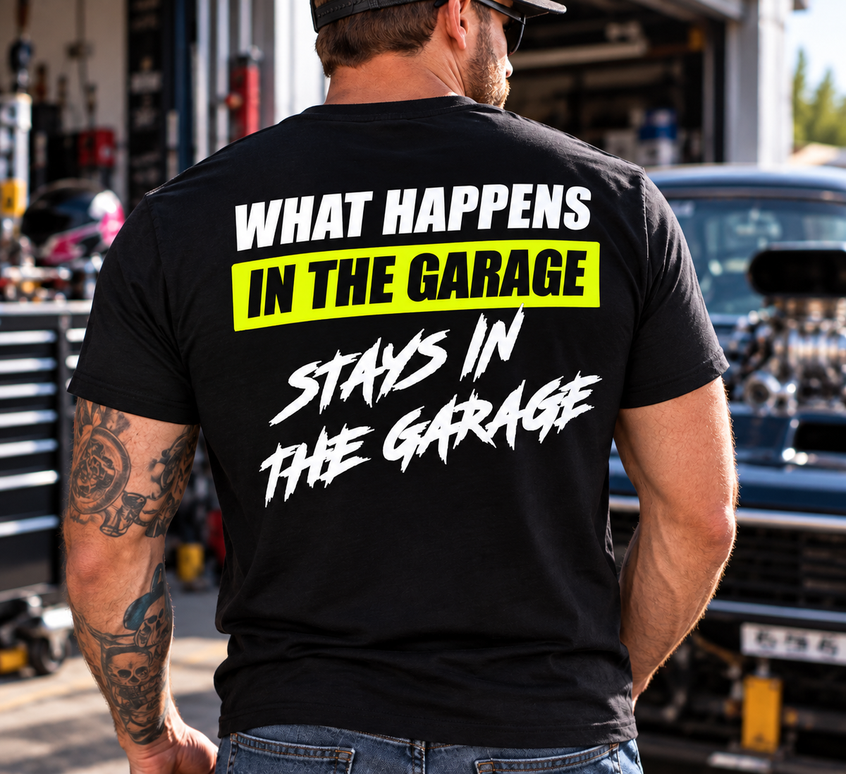 Man wearing a black Car Chix What Happens in the Garage Stays in the Garage T-shirt with white lettering and neon yellow graphic, standing in an automotive garage.