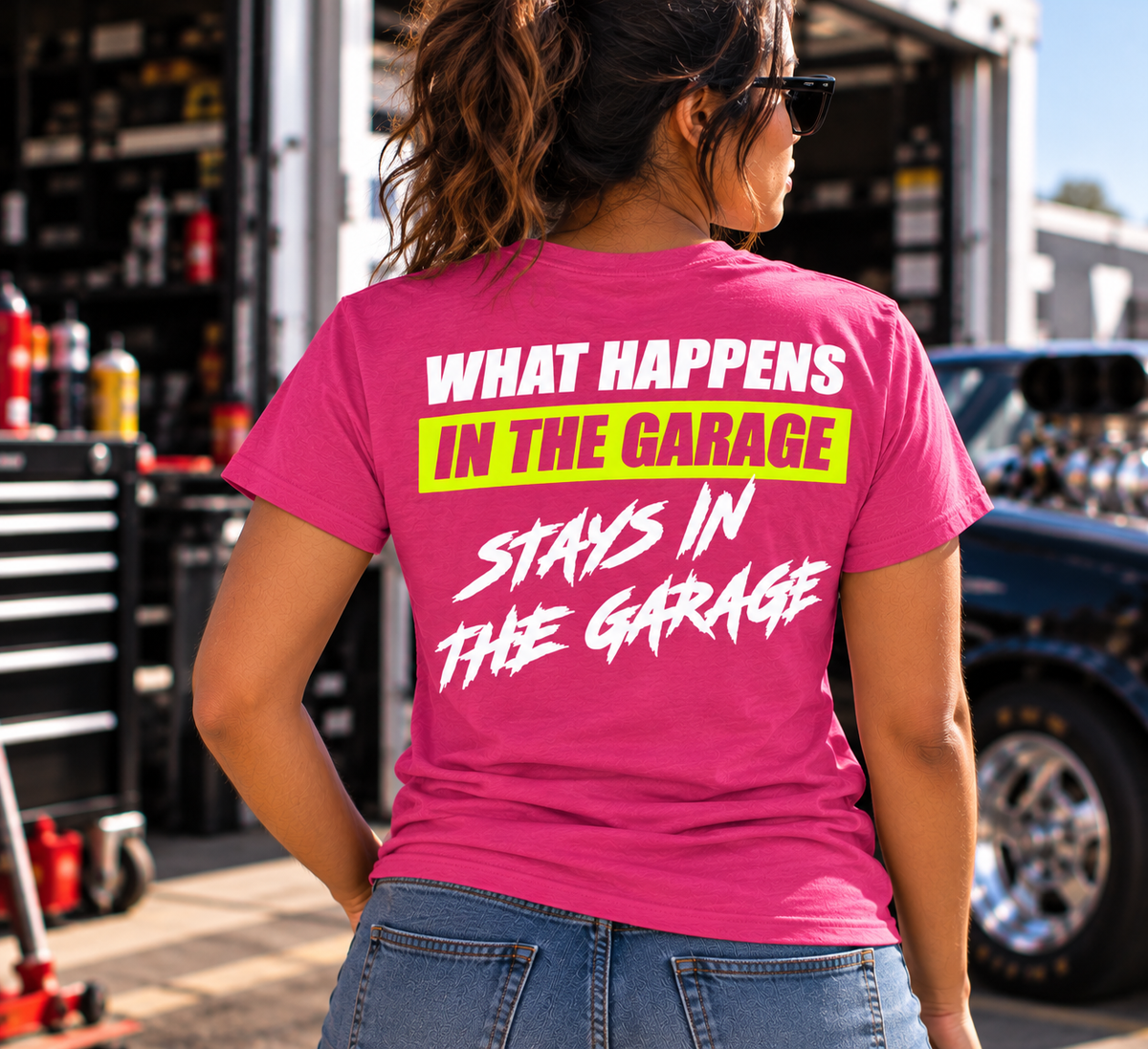 Woman wearing a dark pink Car Chix What Happens in the Garage Stays in the Garage T-shirt with neon yellow and white back graphic at a drag racing garage.