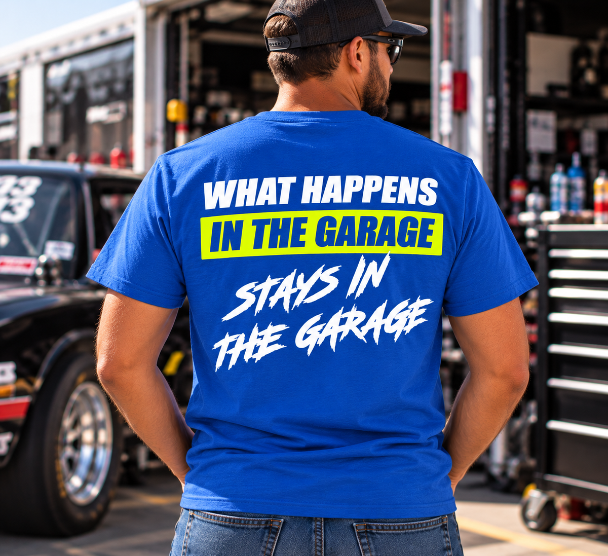 Man wearing a blue Car Chix What Happens in the Garage Stays in the Garage T-shirt featuring a neon yellow and white back print, standing beside a race car.